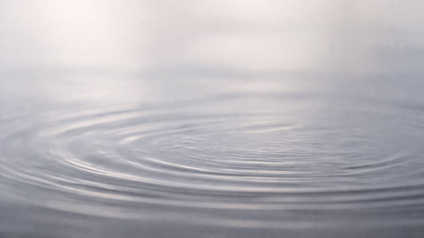 A close-up of a flat water surface with faint, almost imperceptible ripples spreading from a central point, shot in muted natural light against a neutral background.