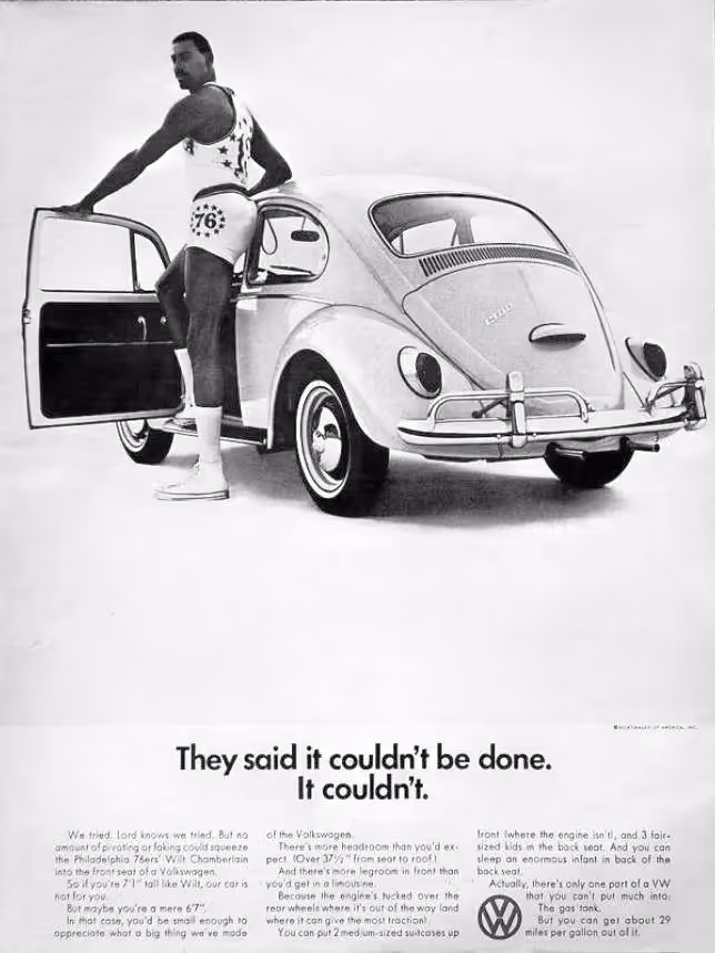 A vintage black and white advertisement showing Wilt Chamberlain, a very tall African American man in a tank top and shorts, standing beside a small Volkswagen Beetle. He has one foot inside the open driver's side door and is leaning on the car's roof, humorously demonstrating his difficulty fitting into the compact car. The headline text reads 'They said it couldn't be done. It couldn't.'