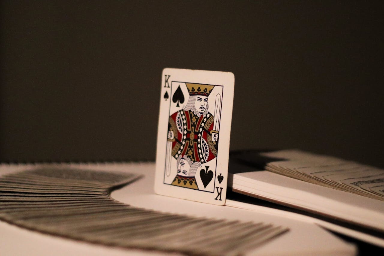King of Spades playing card stands upright on light surface, w/ other playing cards fanned out to its left and a small stack of cards or a book to its right, all against a dark background. Resilience