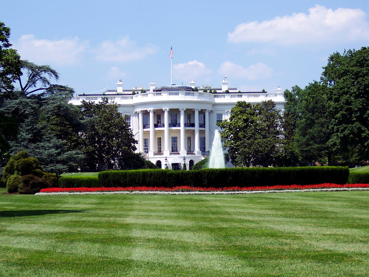 The White House, seen from the South Lawn, featuring its white neoclassical facade with columns, surrounded by lush green trees, a manicured hedge, a vibrant flower bed with red and white blooms, and a tall water fountain, all under a blue sky with scattered white clouds.