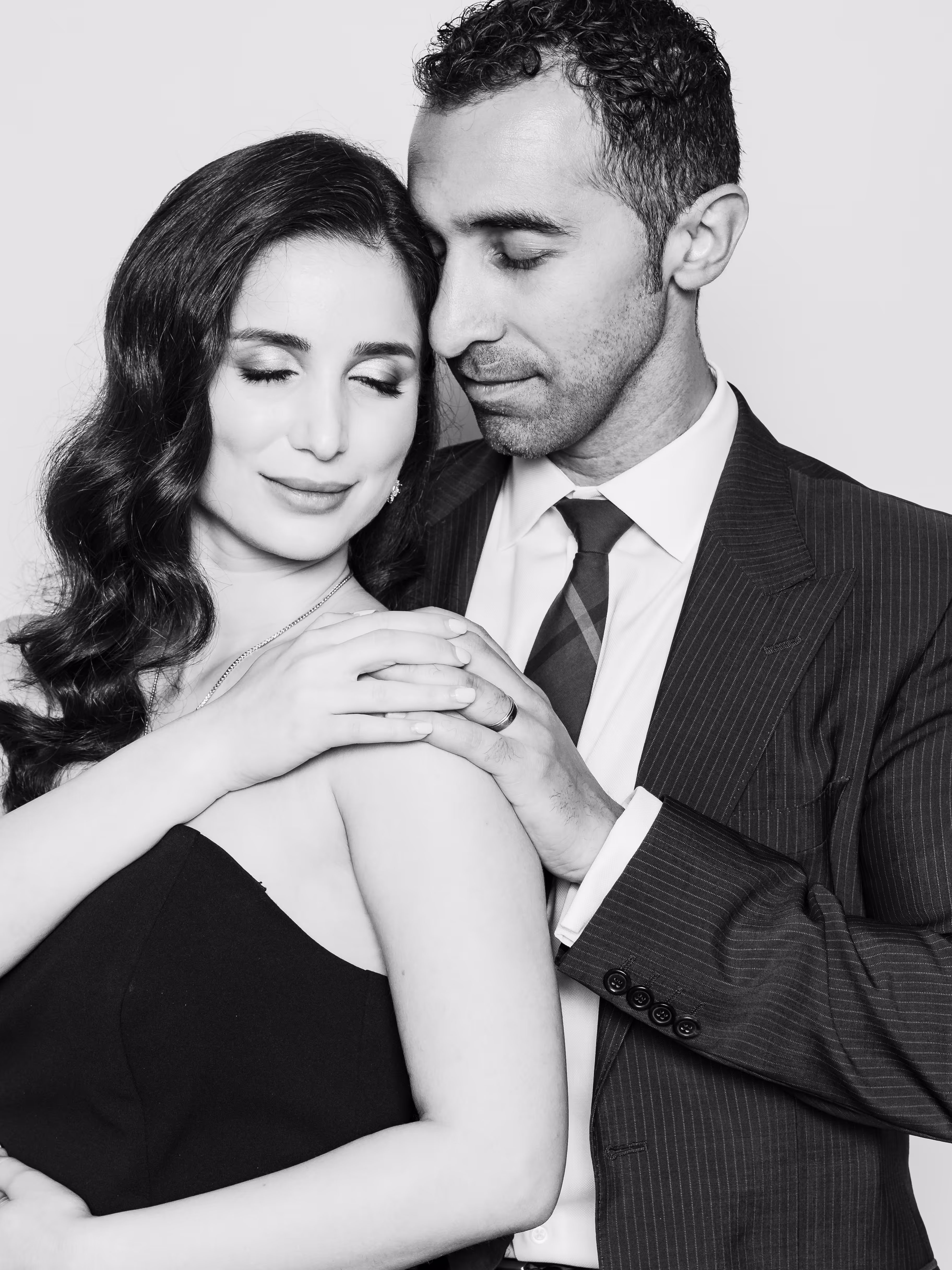 A black and white portrait of a couple embracing intimately. The woman, with dark wavy hair, wears a strapless top and has her eyes closed with a soft smile, her hands gently resting on the man's shoulder. The man, with dark curly hair and a beard, wears a pinstripe suit, white shirt, and striped tie, with his eyes closed, leaning his head close to hers and his hand over hers on his shoulder.